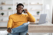 © Prostock-studio - Cheerful young black man sitting at worktable with laptop, looking for job on Internet at home, talking on phone with employer or HR representative, looking at copy space and smiling