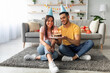© Prostock-studio - A young couple sits on a cozy rug, smiling and celebrating a birthday. The man holds a chocolate cake with lit candles, and they wear party hats in a festive living room.