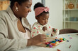 © TrueFrame Collective - Black woman sitting beside Black toddler engaging in creative activity together, toddler holding plastic container while playing with colorful crayons on table