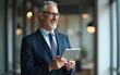 © wang - Happy middle aged business man ceo wearing suit standing in office using digital tablet. Smiling mature businessman professional executive manager looking away. Generative AI. High quality