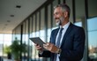 © wu - Happy middle aged business man ceo wearing suit standing in office using digital tablet. Smiling mature businessman professional executive manager looking away thinking working on tech device.