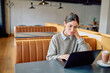 © we.bond.creations - Young woman wearing glasses focusing on her laptop while telecommuting from a modern cafe with a drink on the table