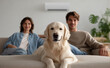 © Jirawatfoto - Happy couple relaxing on sofa with cute golden retriever dog enjoying cool breeze from air conditioner in modern living room, showing content and peaceful expression at home