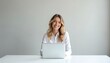 © miss irine - Blonde woman smiles at the camera while sitting at desk. Female uses laptop in bright office. Portrait of happy freelancer looks at display.
