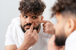 © Maria Vitkovska - Young man examining and squeezing a blemish on his face in the bathroom mirror