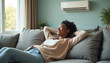© Pete - Happy black woman relaxing on sofa in air conditioned room. Young smiling female enjoys cool fresh air from wall unit. Girl rests comfortably on couch at home escaping summer heat with climate system.