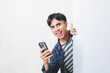 © Muhammad Alimaki - Cheerful Asian man peeking from behind a white wall while holding a smartphone, showing an excited and energetic expression, ideal for concepts of surprise notification, good news, and mobile apps.
