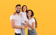 © Prostock-studio - A cheerful family stands together with the father holding their daughter. The mother smiles beside them, all dressed in casual white shirts. The backdrop features a vibrant yellow color.