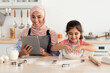 © Prostock-studio - A joyful mother and her daughter are baking together in a sunny kitchen. The daughter is mixing dough while the mother follows a recipe on a tablet. Flour is scattered on the counter.