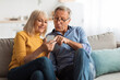 © Prostock-studio - Two adults sit together on a couch in a warm, inviting living room, happily sharing a moment as they look at a smartphone, filled with joy and curiosity.