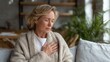 © timitinej - A middle-aged woman is sitting on a couch at home, holding her chest with a worried expression. The soft natural light highlights her concern in this cozy setting