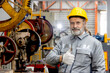 © Stella - Portrait of senior worker wears hard hat, gives thumb up, stands next to machine at manufacturing factory. Elderly engineer technician with safety helmet working with machinery in industrial site.