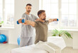 © Studio Romantic - Man patient sitting on the couch in rehabilitation clinic doing sport exercise using dumbbells with nurse. Physiotherapist helping patient in lifting dumbbell. Physical rehabilitation concept.