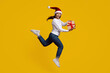 © Prostock-studio - A joyful woman wearing a Santa hat leaps in the air, holding a gift with a red ribbon. The vibrant yellow background enhances her festive spirit, showing excitement for the holidays.