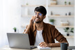 © Prostock-studio - A man with glasses smiles while talking on the phone in a bright and modern workspace. He is sitting at a desk with a laptop and a notebook, looking engaged in conversation.