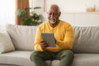 © Prostock-studio - A senior man smiles as he interacts with a tablet in a bright living room. He is comfortably seated on a soft sofa, enjoying his time while exploring content on the device.