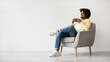 © Prostock-studio - A happy young African American man relaxes in a chair, sipping coffee. He smiles while looking off to the side, enjoying a moment of calm in a simple setting at home.