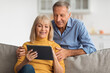 © Prostock-studio - A happy older couple sits closely on a sofa, smiling as they look at a tablet together. They are in a bright living room filled with warmth and comfort.