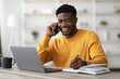 © Prostock-studio - Happy african american young man freelancer or businessman working from home, sitting at table in front of modern laptop, having phone conversation, taking notes and smiling, copy space