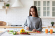 © BestForYou - Woman in a gray sweater is standing in a kitchen with fresh vegetables