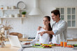 © BestForYou - Woman in a white long-sleeve shirt pointing at a laptop, standing in a kitchen with a man in a light blue shirt behind her.