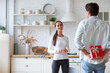 © BestForYou - Woman in a white long-sleeve shirt clapping her hands, standing in a kitchen with a man in a light blue shirt behind her. The man is holding a red gift box with a white ribbon.