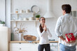 © BestForYou - Woman in a white long-sleeve shirt clapping her hands, standing in a kitchen with a man in a light blue shirt behind her. The man is holding a red gift box with a white ribbon.