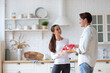 © BestForYou - Woman in a white long-sleeve shirt clapping her hands, standing in a kitchen with a man in a light blue shirt behind her. The man is holding a red gift box with a white ribbon.