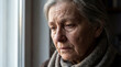 © MYKHAILO KUSHEI - An older woman stands near a bright window, her face etched with sadness as a tear escapes her eye, reflecting deep emotional pain and quiet solitude.