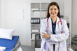 © Wasan - Female doctor wearing a white coat and stethoscope smiles confidently with her arms crossed, standing in hospital room with a patient bed and laptop in the background.