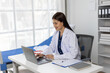 © Wasan - Young female doctor smiling while working on a laptop in medical office, with a stethoscope and medical records on the desk and a hospital bed nearby.