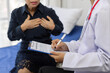 © Wasan - Senior woman sitting on a hospital bed, discussing chest pain symptoms with a doctor, who is taking notes on a clipboard during their consultation in the examination room.