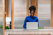 © artitwpd - An African woman in a blue top works on her laptop at a modern reception desk, surrounded by wooden and glass partitions, reflecting a professional, organized office environment.