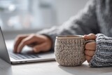 Woman in warm sweater holding coffee mug while working on laptop. Cozy office space for comfortable remote work or studying.