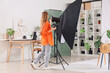 © Pixel-Shot - Young female photographer with houseplants, photo camera and lighting equipment preparing scene for shooting in studio