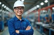 © miss irine - Asian man in hard hat smiles in factory. Worker with glasses wears blue shirt with arms crossed. Industrial warehouse setting with machines and lights.