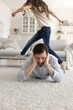 © fizkes - Parenting is hard work. Exhausted upset mid aged dad lying on warm living room floor hug head in despair unable to deal with active energetic small daughter jumping running creating noisy chaos around