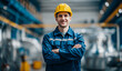 © andyaziz6 - Smiling young male industrial worker wearing a bright yellow safety helmet and blue reflective uniform standing confidently with arms crossed inside a modern factory environment