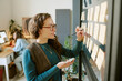 © AnnaStills - Caucasian young adult woman writing on sticky notes attached to glass wall, holding marker and eraser, working in modern office while another young adult woman works at computer in background