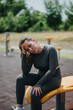 © qunica.com - A woman in grey athletic wear sits on an outdoor bench, appearing fatigued post-workout. The background features blurred greenery, enhancing the serene yet determined focus of the fitness setting.