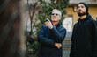 © qunica.com - An elderly man in a quilted jacket points as he speaks to a younger man wearing a beanie. They stand outside a building with greenery, sharing a moment of guidance and connection.
