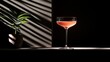 © mypix - Elegant cocktail in a stemmed glass sits on a shelf with a potted plant, shadowed by light. Dark backdrop
