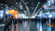 © wolfhound911 - A blurred scene of a bustling trade fair or expo, with large crowds exploring booths and product displays.