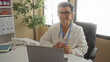 © Krakenimages.com - Senior woman doctor with grey hair in a clinic office holding a stethoscope smiling at her desk surrounded by office equipment and a plant.