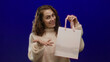 © Krakenimages.com - Woman happily holding shopping bag against isolated blue background wearing cozy sweater with brunette hair smiling and showcasing casual fashion style