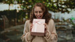 © Krakenimages.com - Woman holding pink gift box with ribbon outdoor in city street, smiling happily wearing glasses and sweater, surrounded by blurred urban background.