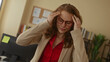 © Krakenimages.com - Young woman in glasses holding her head, appearing stressed in an office with a computer and bookshelf behind her, suggesting a challenging work environment indoors.