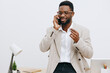 © SHOTPRIME STUDIO - Professional shot of a smiling man with dark skin and glasses talking on a mobile phone while standing in an office environment, wearing a light blazer and white shirt, with a desk, laptop, and plant