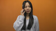© Krakenimages.com - Woman smiling while talking on mobile against vibrant orange background, highlighting youthful asian features and casual attire in an isolated setting.