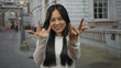 © Krakenimages.com - Young chinese woman making rock gesture with both hands outdoors on a street with an urban background, wearing a white shirt and smiling confidently at the camera.